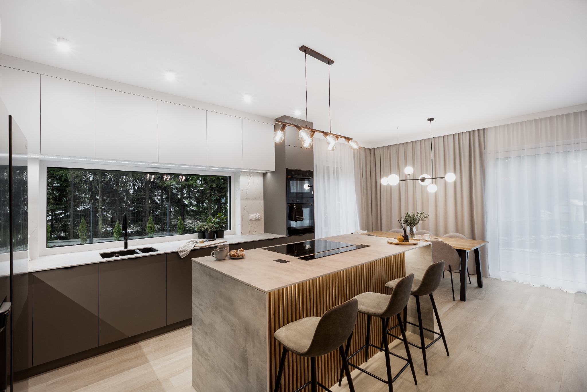 Nowoczesna kuchnia z wyspą wykończoną drewnianymi lamelami, minimalistyczne szafki, duże okno z widokiem na zieleń, oświetlenie punktowe i dekoracyjne lampy wiszące.