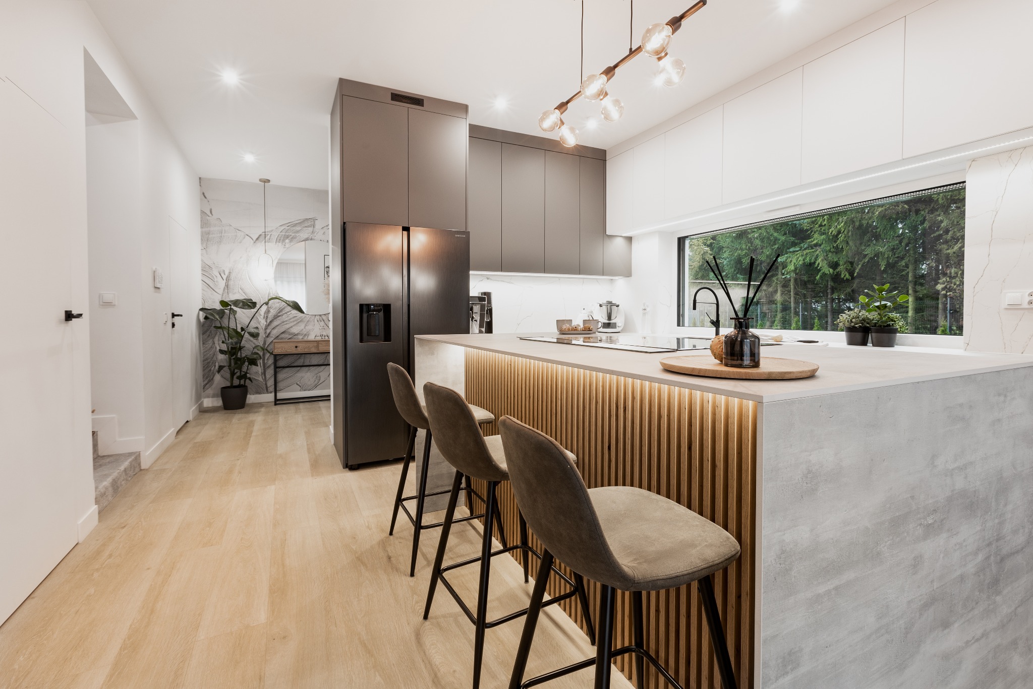 Nowoczesna kuchnia z wyspą, drewnianymi lamelami podświetlonymi LED, szarymi szafkami i czarną lodówką Samsung, widok z perspektywy podłogi.