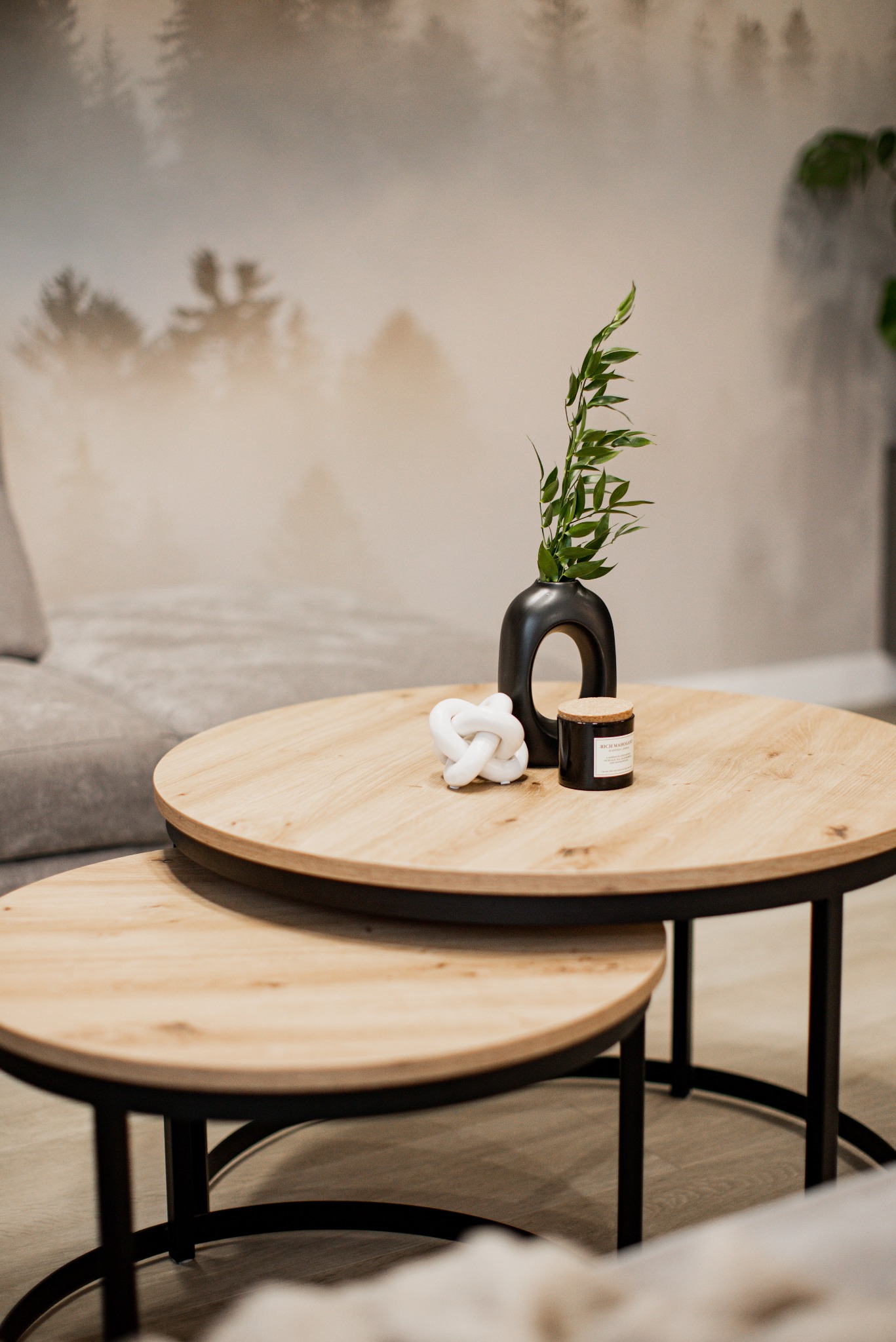 Zestaw dwóch okrągłych stolików kawowych z drewnianymi blatami i czarnymi metalowymi nogami, na większym stoliku czarny wazon z zielonymi gałązkami, biała dekoracja w kształcie węzła i czarna...