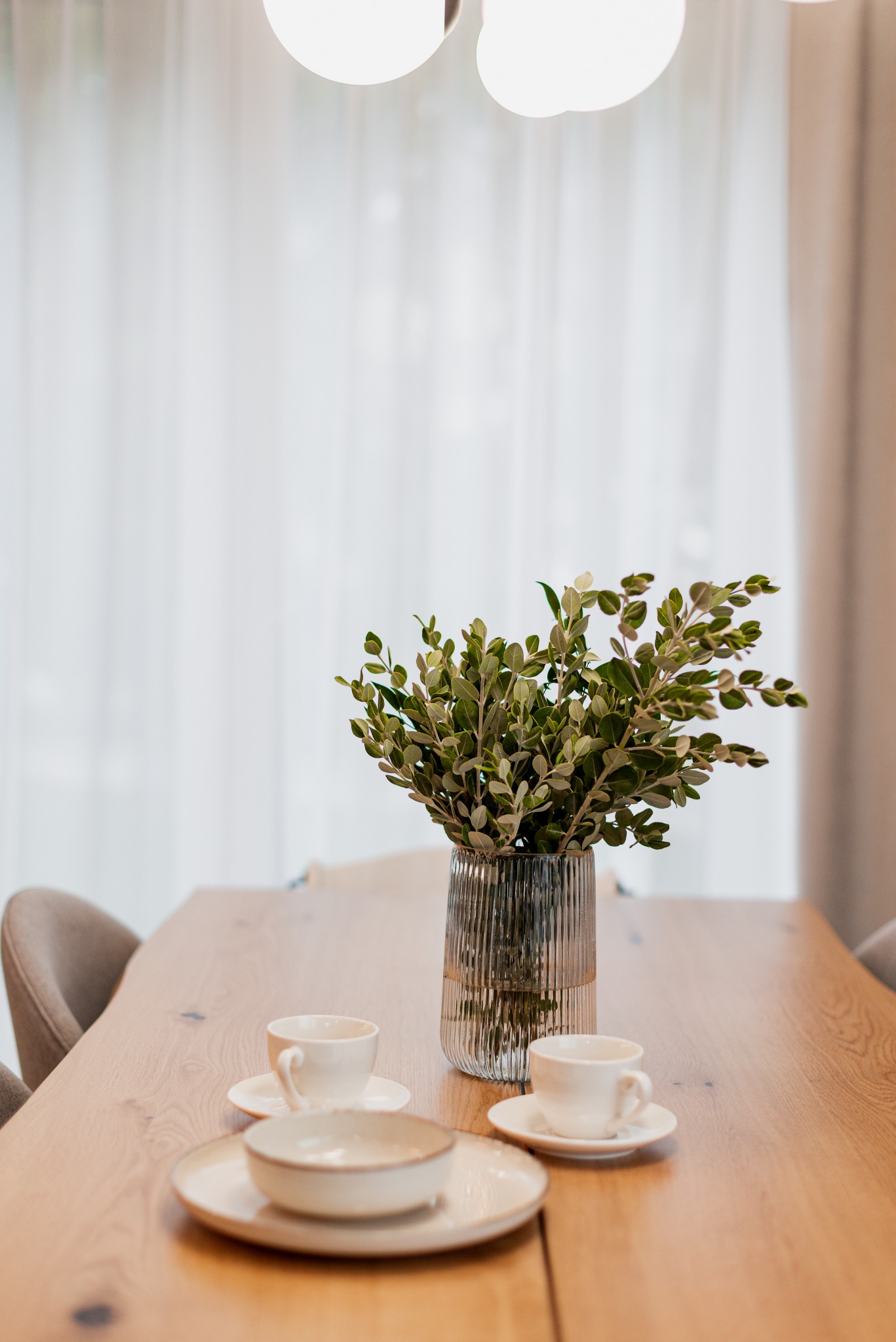 Drewniany stół jadalny z blatem z widocznymi słojami, nakryty białymi filiżankami i talerzami, dekorowany szklanym wazonem z zielonymi gałązkami, oświetlony lampą sufitową na tle białej zasłony.