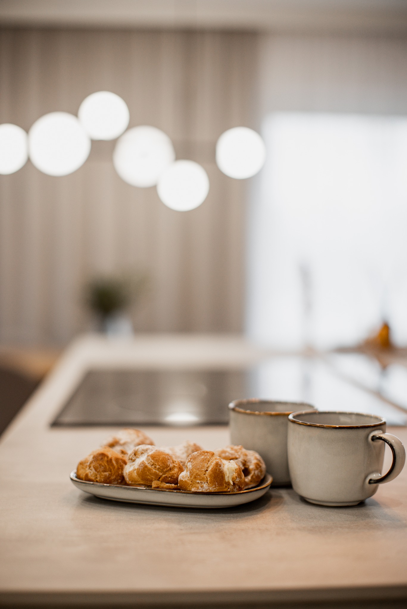 Jasna kuchnia z blatem, na którym leżą pączki na talerzyku i dwie ceramiczne filiżanki z brązowymi obwódkami.
