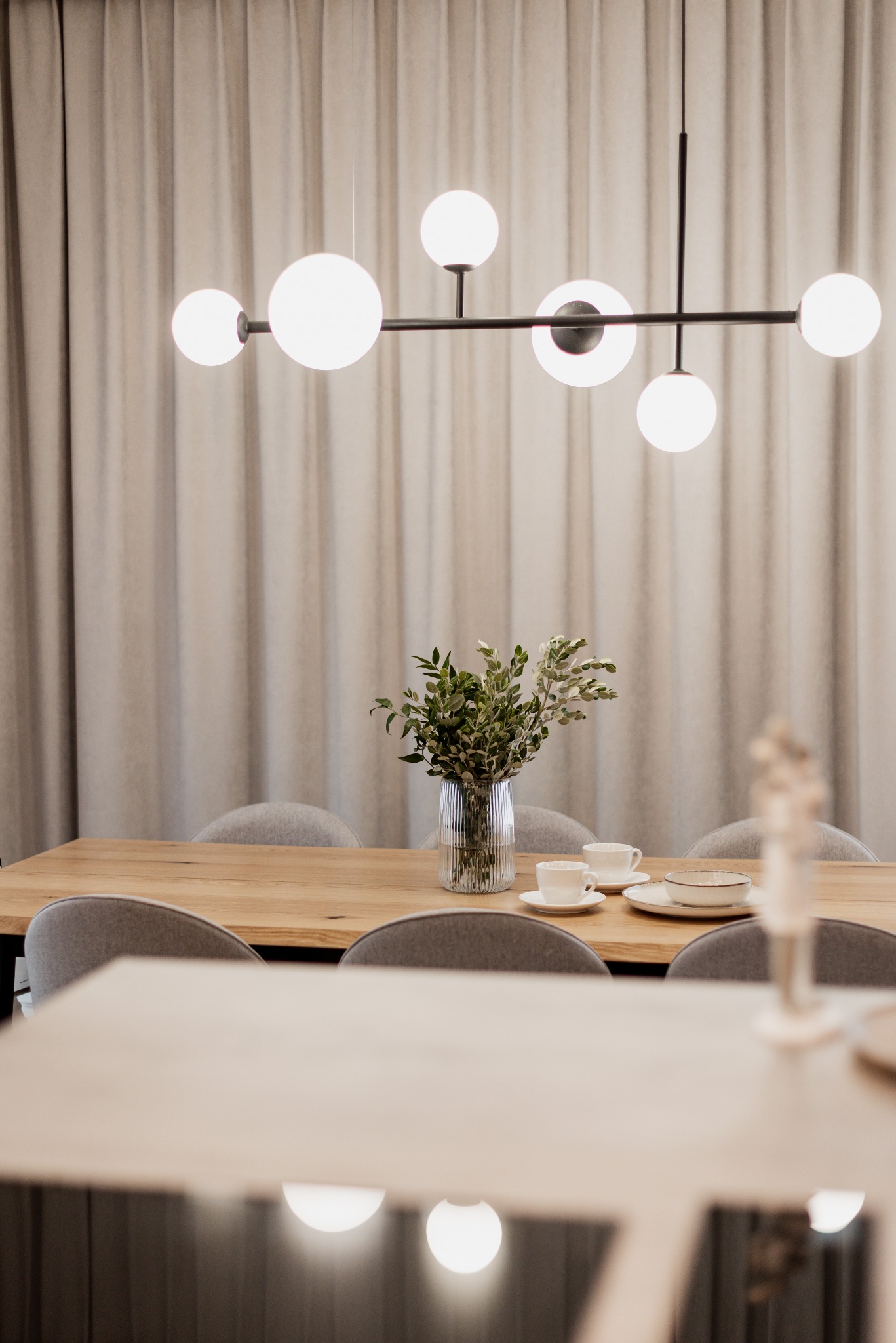 Jasny pokój jadalny z drewnianym stołem, szarymi krzesłami, nowoczesną lampą wiszącą z białymi kulami i wazonem z zielonymi gałązkami na stole, nakryty do posiłku.