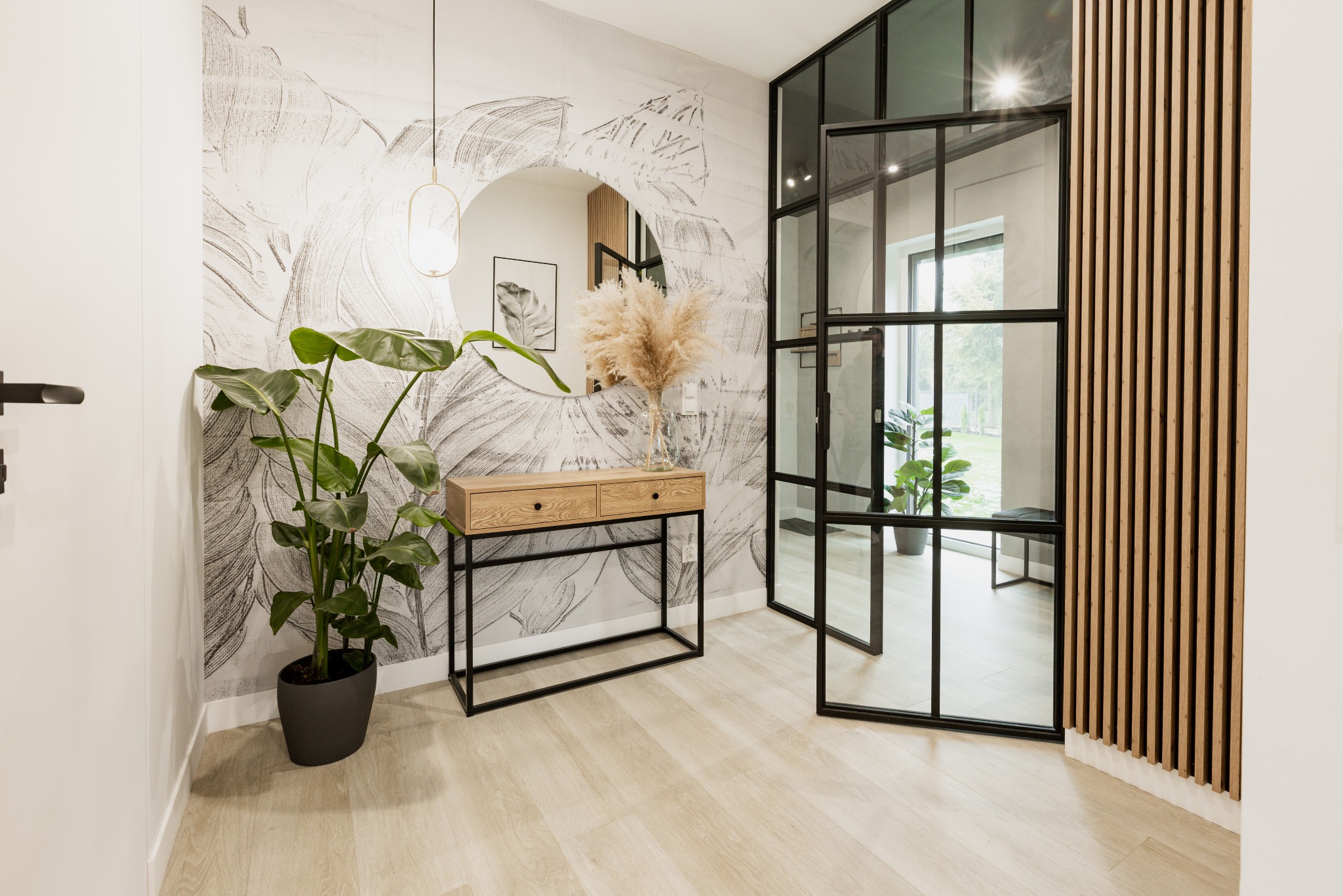 Stylowy przedpokój w Bydgoszczy z konsolą z dwiema szufladami, okrągłym lustrem i dekoracyjną ścianą z motywem roślinnym. W tle widoczne drzwi w stylu loftowym z czarnymi szprosami i drewniane lamele.
