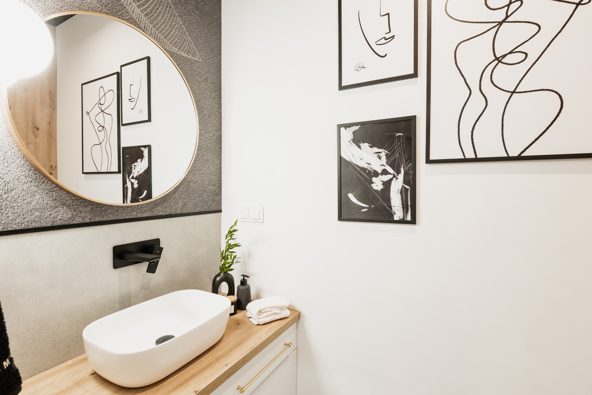 Nowoczesna łazienka z owalnym lustrem w złotej ramie, umywalką nablatową, czarną armaturą, drewnianym blatem i minimalistycznymi grafikami na ścianie.