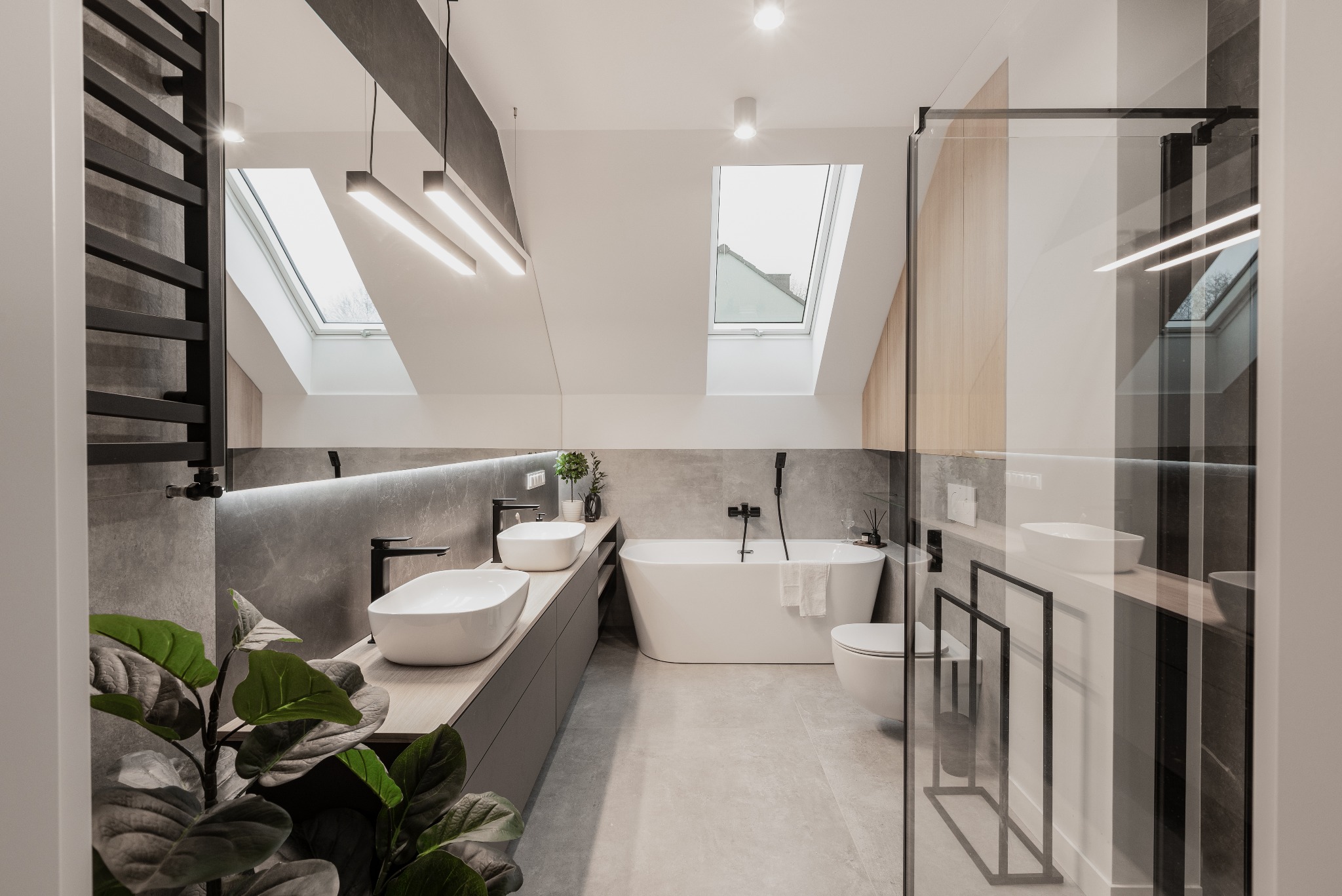 Nowoczesna łazienka na poddaszu ze skośnym sufitem i oknami dachowymi, wyposażona w dwie białe umywalki nablatowe, wolnostojącą wannę, szklaną kabinę prysznicową i grzejnik drabinkowy w kolorze...