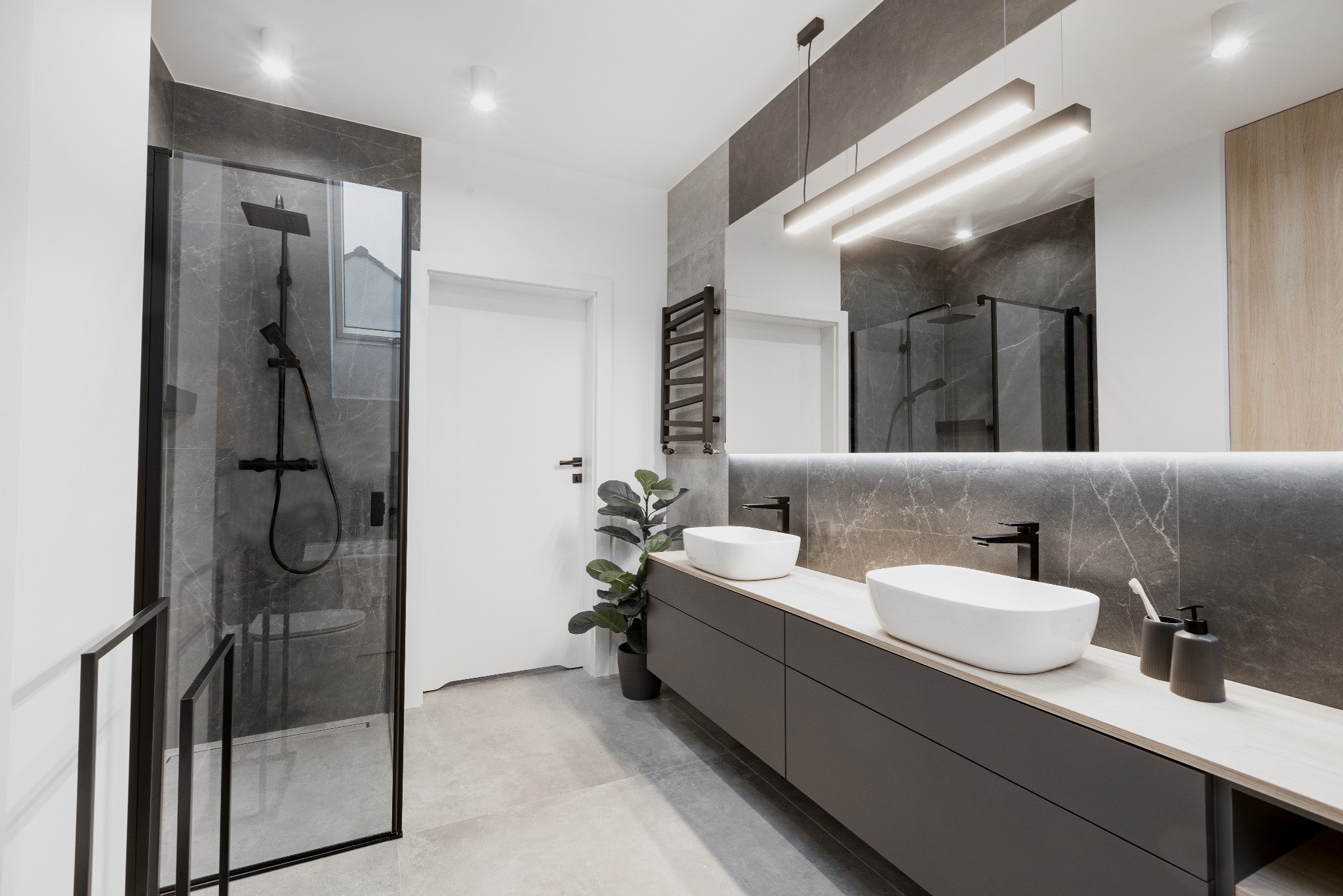 Nowoczesna łazienka z szarą armaturą, kabiną prysznicową z czarnymi ramami, dwiema białymi umywalkami na szarej szafce z drewnianym blatem, podświetlanym lustrem i grzejnikiem drabinkowym.