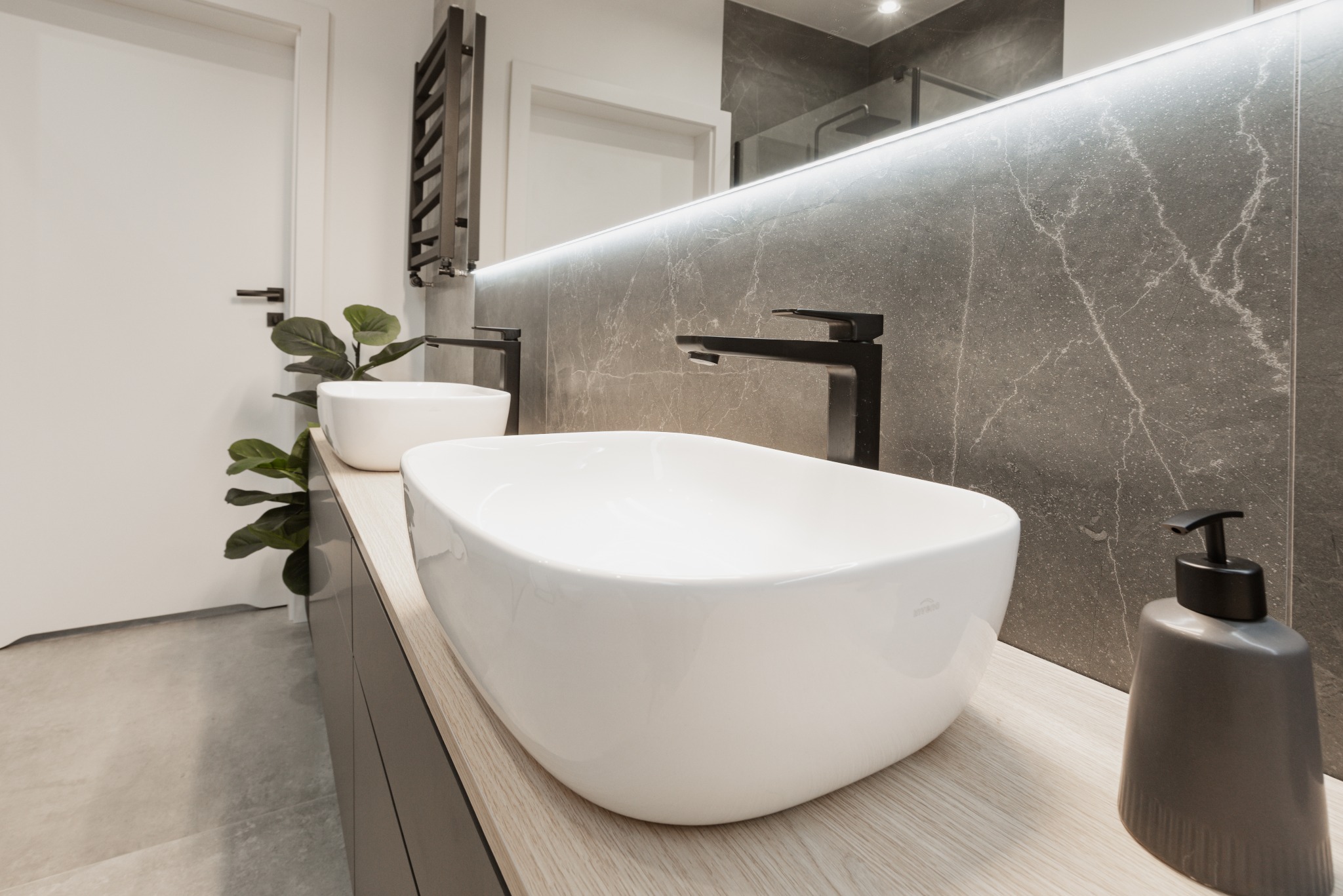 Nowoczesna łazienka z dwoma białymi umywalkami nablatowymi, czarną armaturą, szarą marmurową ścianą z podświetlanym lustrem, grzejnikiem drabinkowym i rośliną doniczkową