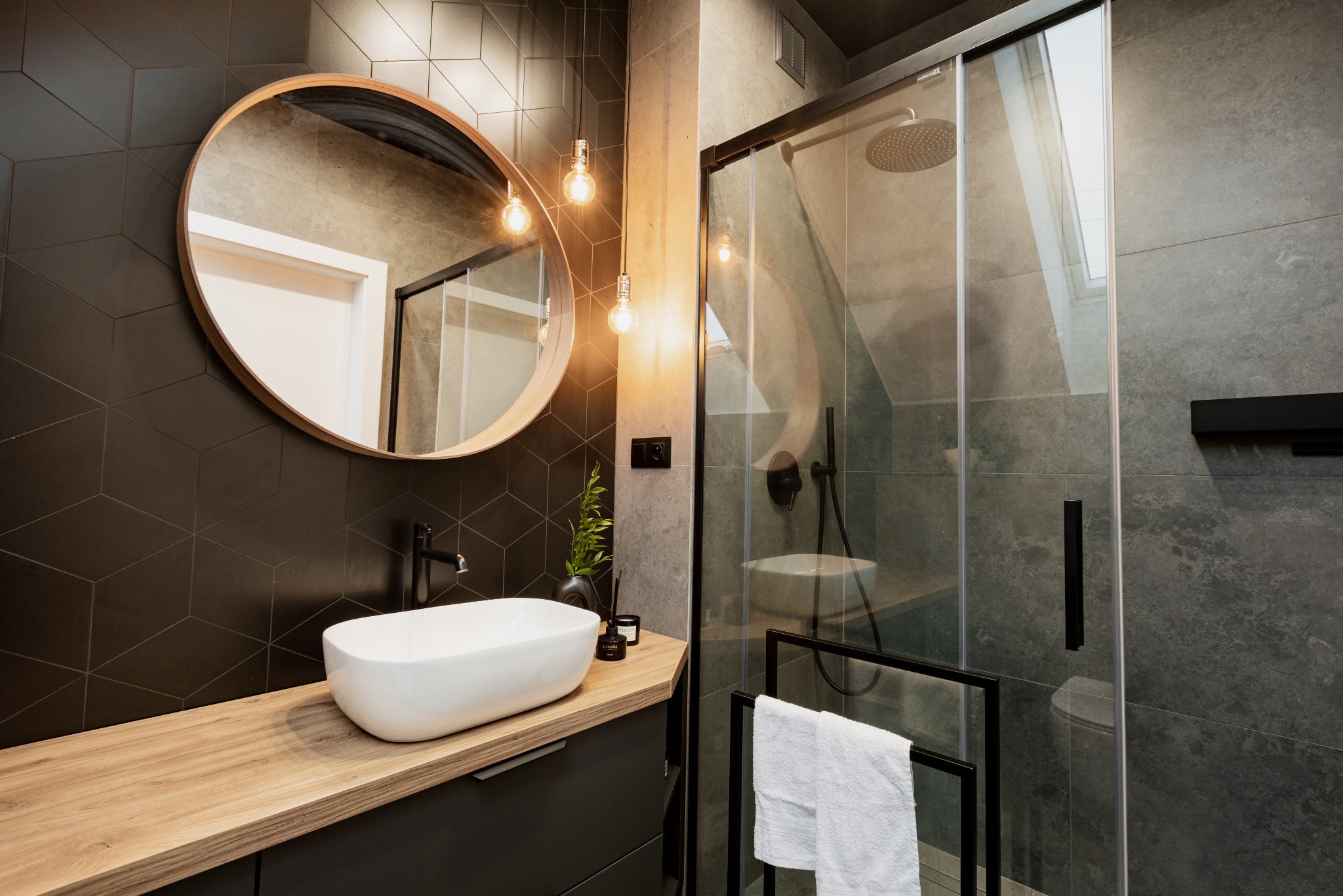 Nowoczesna łazienka z umywalką nablatową, okrągłym lustrem w drewnianej ramie, kabiną prysznicową ze szklanymi drzwiami i czarną armaturą, oświetlona wiszącymi żarówkami edisona.