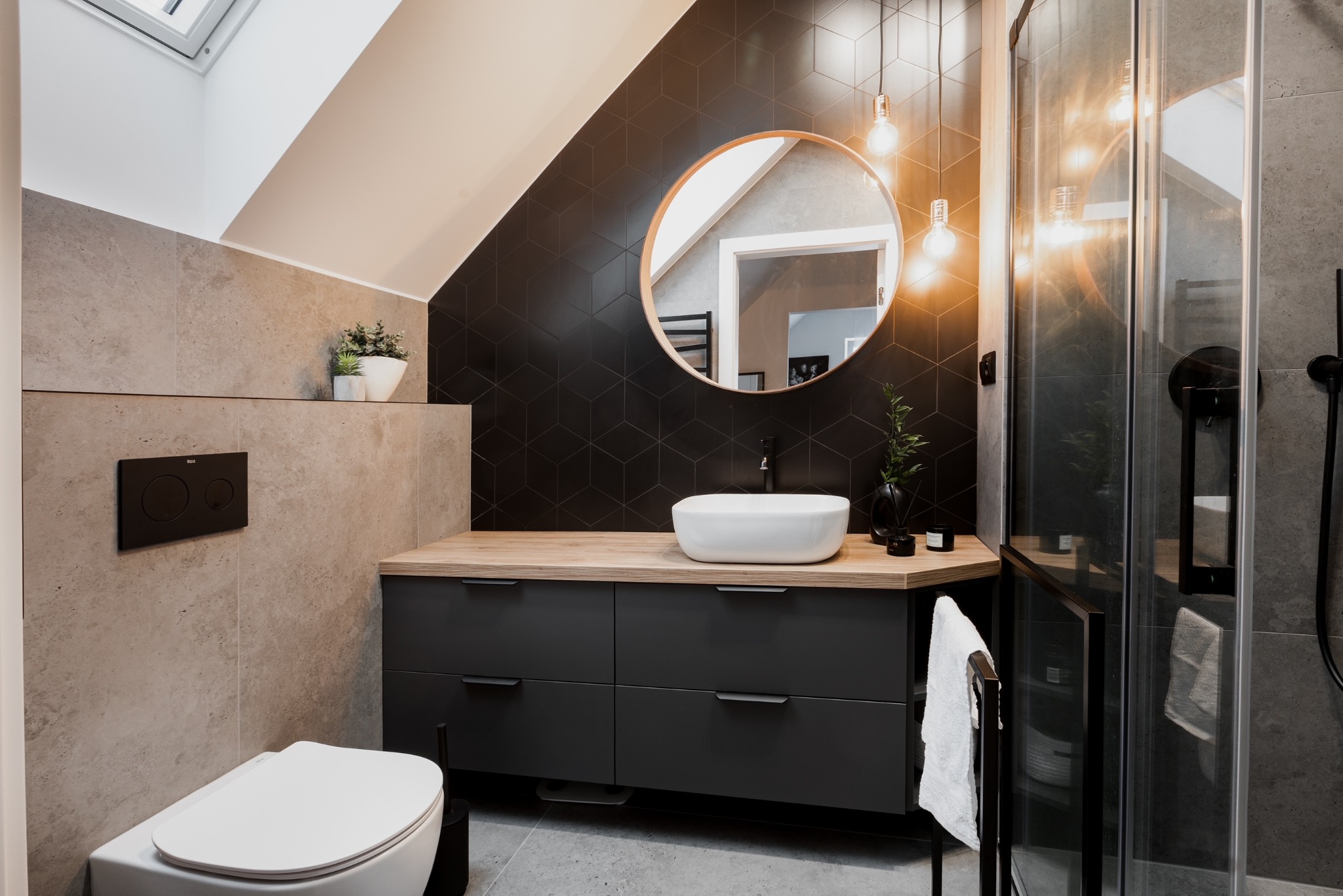 Nowoczesna łazienka na poddaszu z czarną ścianą z heksagonów, okrągłym lustrem, drewnianym blatem i szarą szafką pod umywalką oraz kabiną prysznicową ze szklanymi drzwiami.