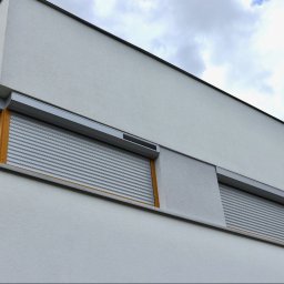 ARESA EU Sp. z o.o. - Dwa okna z szarymi, aluminiowymi roletami zewnętrznymi na białej fasadzie nowoczesnego domu, widziane pod kątem z dołu. Częściowo widoczne błękitne niebo.