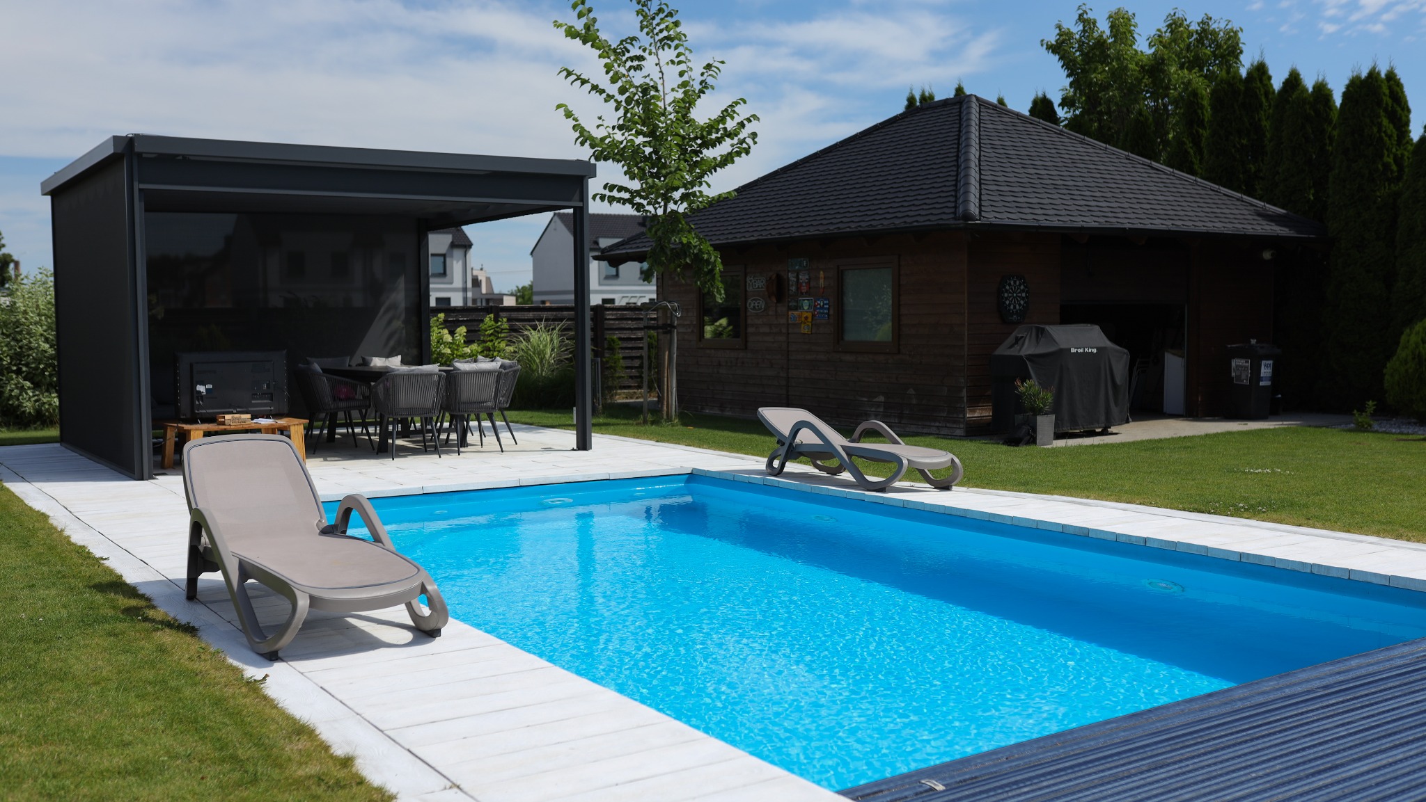 Nowoczesny basen z turkusową wodą, otoczony białymi płytami, z szarą pergolą z zasłoniętymi bokami, leżakami i małym domkiem narzędziowym w tle.