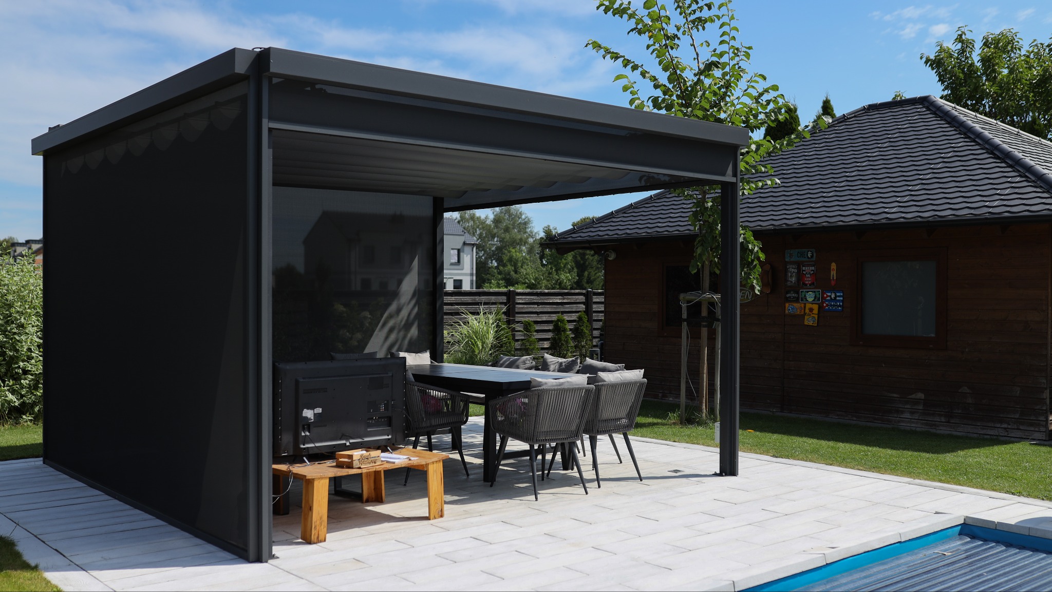 Nowoczesna pergola z czarnymi roletami bocznymi, wyposażona w stół z krzesłami i telewizor, na tle zadbanego ogrodu z drewnianym domkiem.