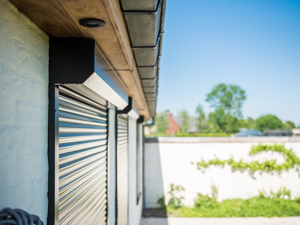 Zamknięte, czarne rolety zewnętrzne na ścianie domu z fakturą, widok z boku w słoneczny dzień.