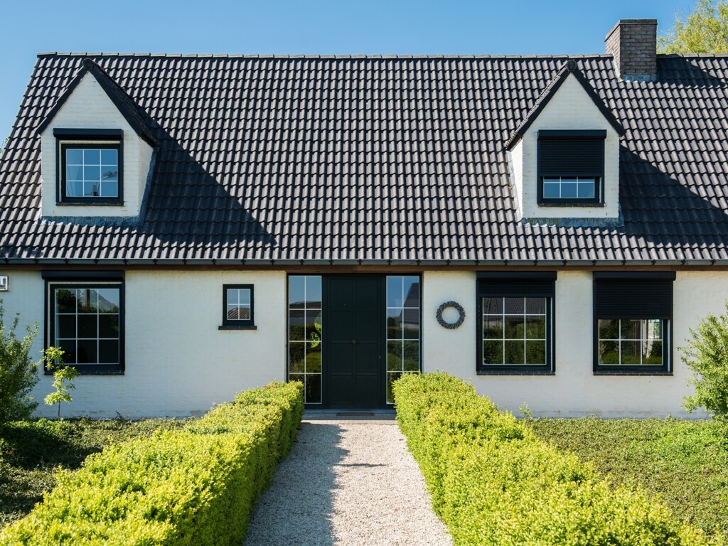 Dom jednorodzinny z białymi ścianami i czarnymi roletami zewnętrznymi, widok z ogrodu z równo przyciętym żywopłotem wzdłuż ścieżki.