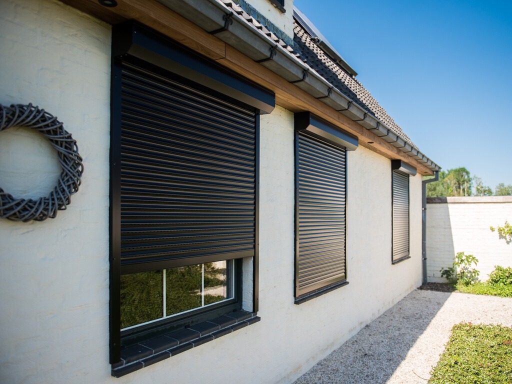 Elewacja domu z trzema oknami zabezpieczonymi czarnymi roletami zewnętrznymi, widoczny dekoracyjny wieniec na ścianie i fragment ogrodu.