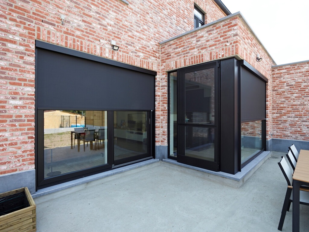 Zewnętrzne, zaciemniające rolety w kolorze antracytowym zamontowane na oknach i szklanych drzwiach tarasowych domu z czerwonej cegły.