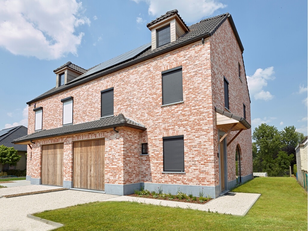 Dwukondygnacyjny dom z cegły klinkierowej z zamontowanymi roletami zewnętrznymi w kolorze antracytowym i białym, widoczne panele słoneczne na dachu.