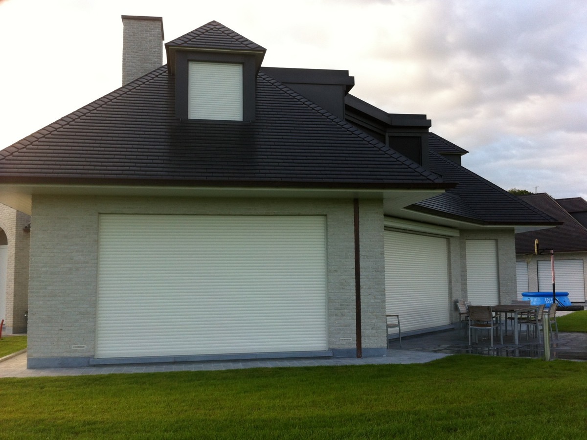 Dom z szarej cegły z trzema białymi roletami zewnętrznymi, stolik i krzesła na zewnątrz.