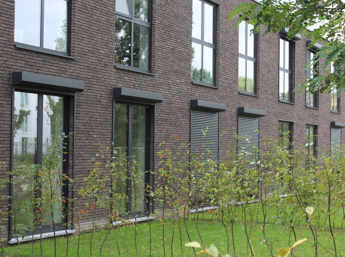 Elewacja budynku z brązowej cegły z oknami wyposażonymi w szare zewnętrzne osłony okienne, część z nich jest zwinięta, a część zasłonięta, widoczne młode krzewy przed budynkiem.