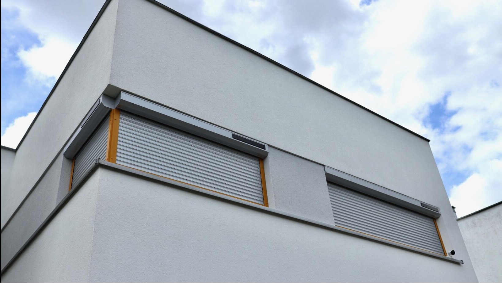 Dwa okna z szarymi, aluminiowymi roletami zewnętrznymi na białej fasadzie nowoczesnego domu, widziane pod kątem z dołu. Częściowo widoczne błękitne niebo.