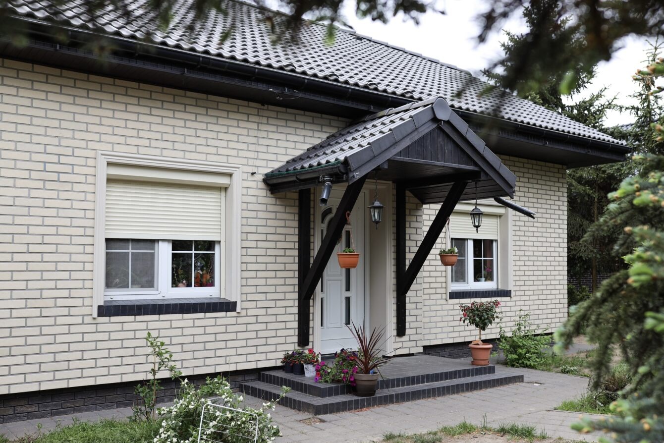 Dom z jasnej cegły z beżowymi roletami zewnętrznymi na oknach. Wejście z zadaszeniem i wiszącymi doniczkami. Widok z poziomu ogrodu.