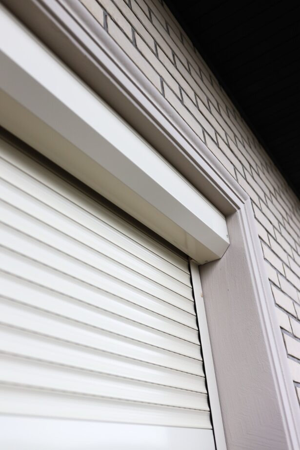 Zamknięta, kremowa osłona okienna na tle jasnej, ceglanej ściany. Widok z dołu, ukazujący detale konstrukcji i fakturę materiałów.