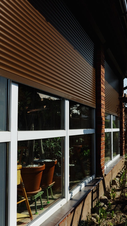 Zewnętrzne rolety w kolorze brązowym, zamontowane na oknach budynku z ceglaną elewacją. Widoczne rośliny w donicach za szybą i fragment ogrodu.