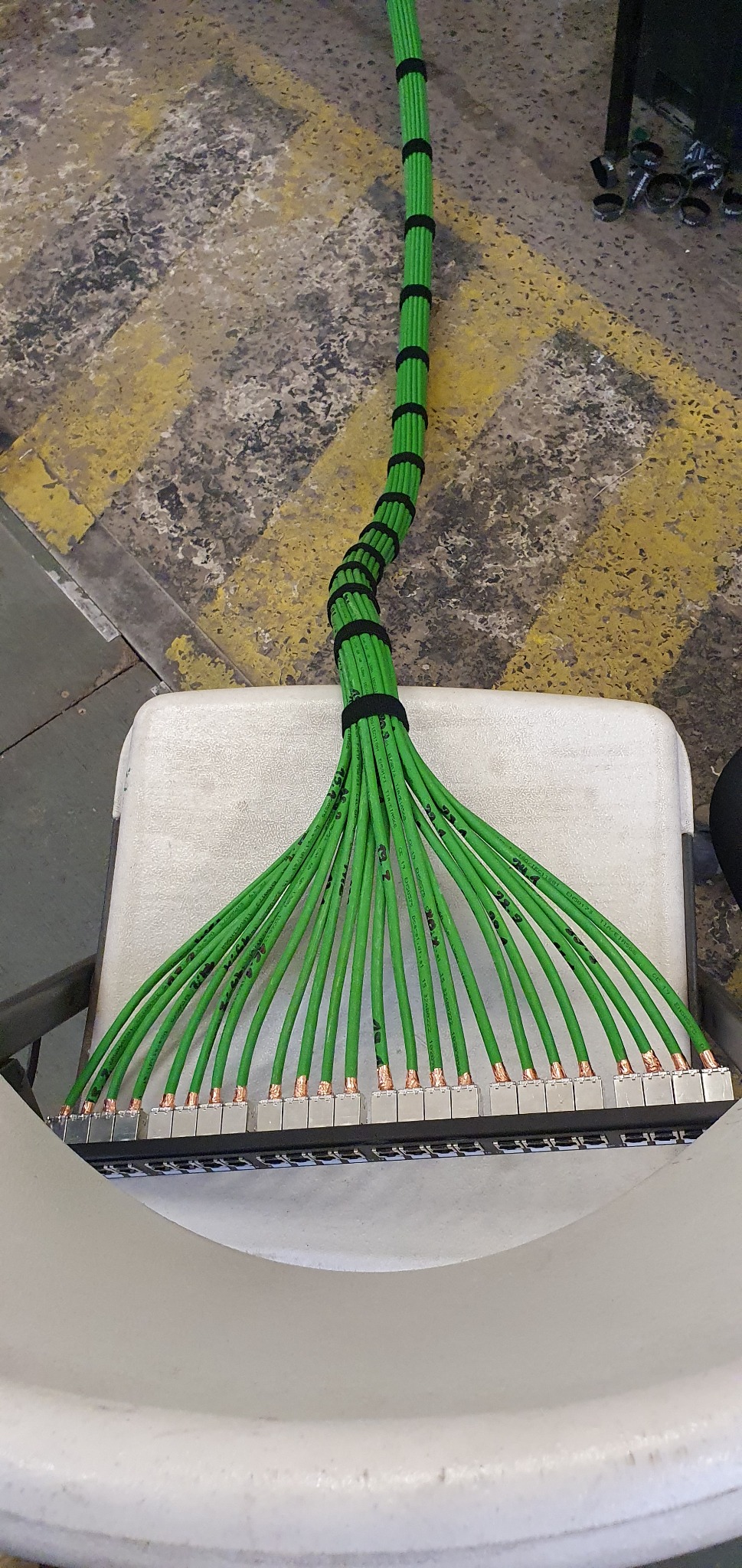 Zestaw podłączonych, oznaczonych zielonych kabli sieciowych UTP z końcówkami RJ45, zebranych w wiązkę czarnymi opaskami zaciskowymi, leżących na białym krześle na tle podłogi z żółtymi pasami.