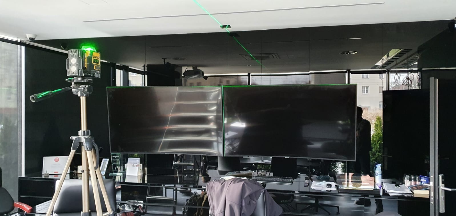 Laser krzyżowy ustawiony na statywie w nowoczesnym biurze z czarnymi ścianami i dwoma dużymi monitorami, emitujący zielone linie na suficie i ekranach.