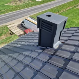 Nowy dach z szarej dachówki ceramicznej z czarnym kominem wentylacyjnym, widok z góry, w tle budynek w trakcie budowy i samochód dostawczy z deskami na dachu.