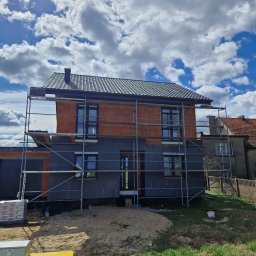 USŁUGI OGÓLNOBUDOWLANE Łukasz Poczontek - Dom w trakcie budowy, z nowym, ciemnym dachem i rusztowaniem, ocieplany styropianem, na tle błękitnego nieba z chmurami.