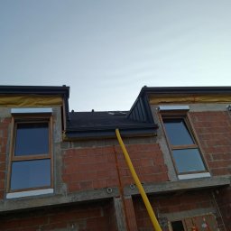 USŁUGI OGÓLNOBUDOWLANE Łukasz Poczontek - Montaż dachu z blachy na nowym budynku z cegły, widoczne okna dachowe i żółty przewód wentylacyjny. Dach w kolorze grafitowym, wykończenia blacharskie.