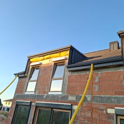 USŁUGI OGÓLNOBUDOWLANE Łukasz Poczontek - Dom w trakcie budowy: ceglana ściana z oknami, fragment dachu z czarną blachą, żółte rury wentylacyjne. Widok na błękitne niebo.