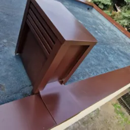 Brązowy kominek wentylacyjny na dachu pokrytym papą, z widocznymi obróbkami blacharskimi i fragmentem zabudowy mieszkalnej w tle.