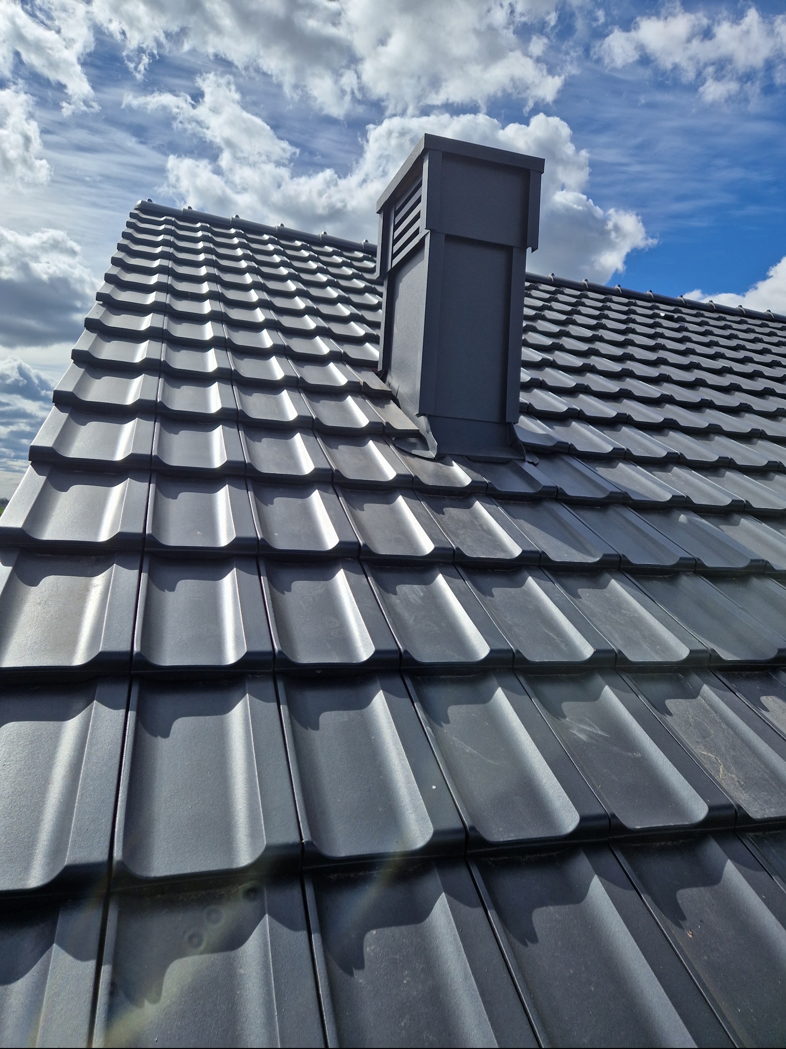 Grafitowy dach z blachodachówki modułowej z kominem wentylacyjnym na tle błękitnego nieba z chmurami.