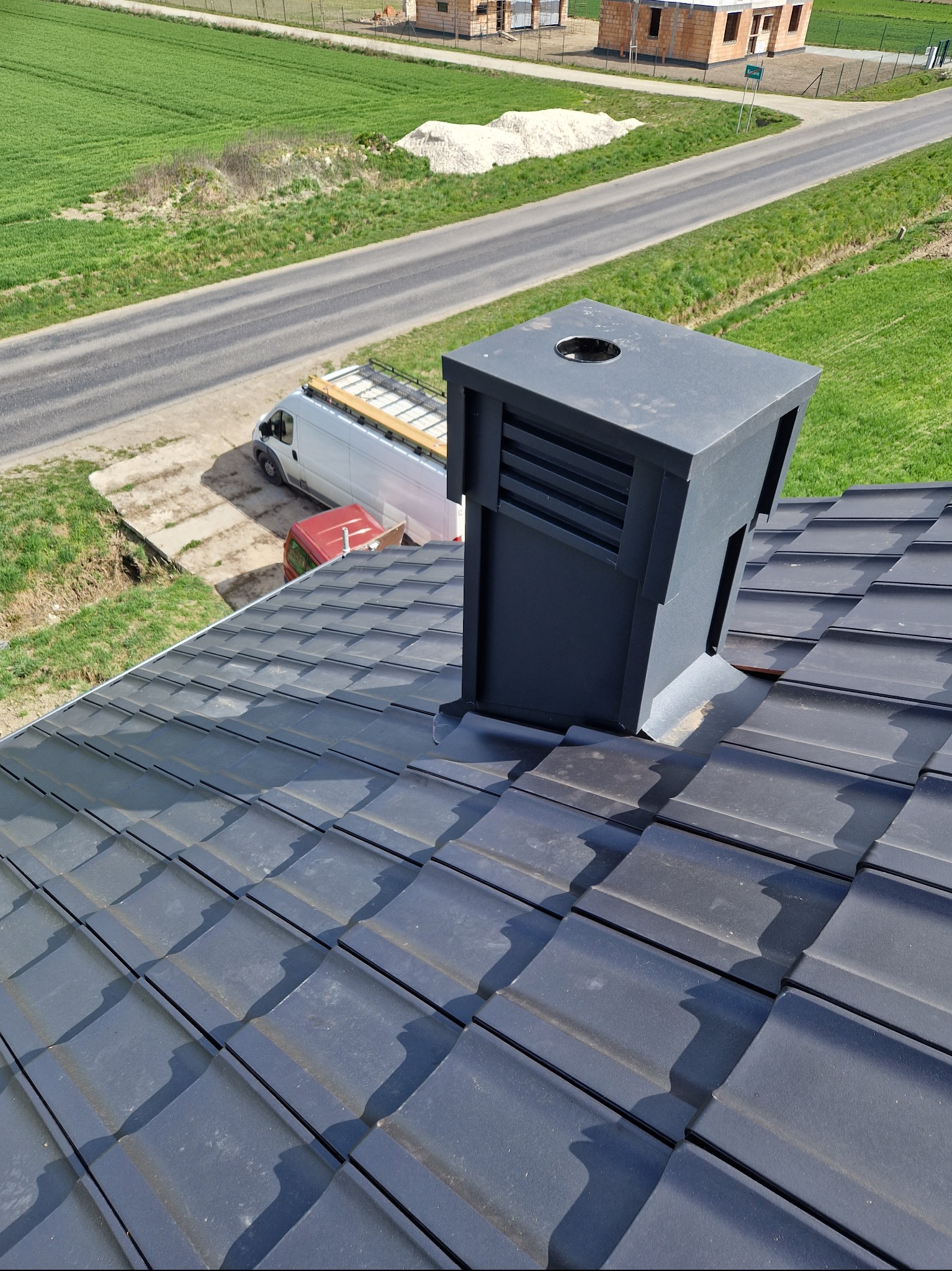 Nowy dach z szarej dachówki ceramicznej z czarnym kominem wentylacyjnym, widok z góry, w tle budynek w trakcie budowy i samochód dostawczy z deskami na dachu.