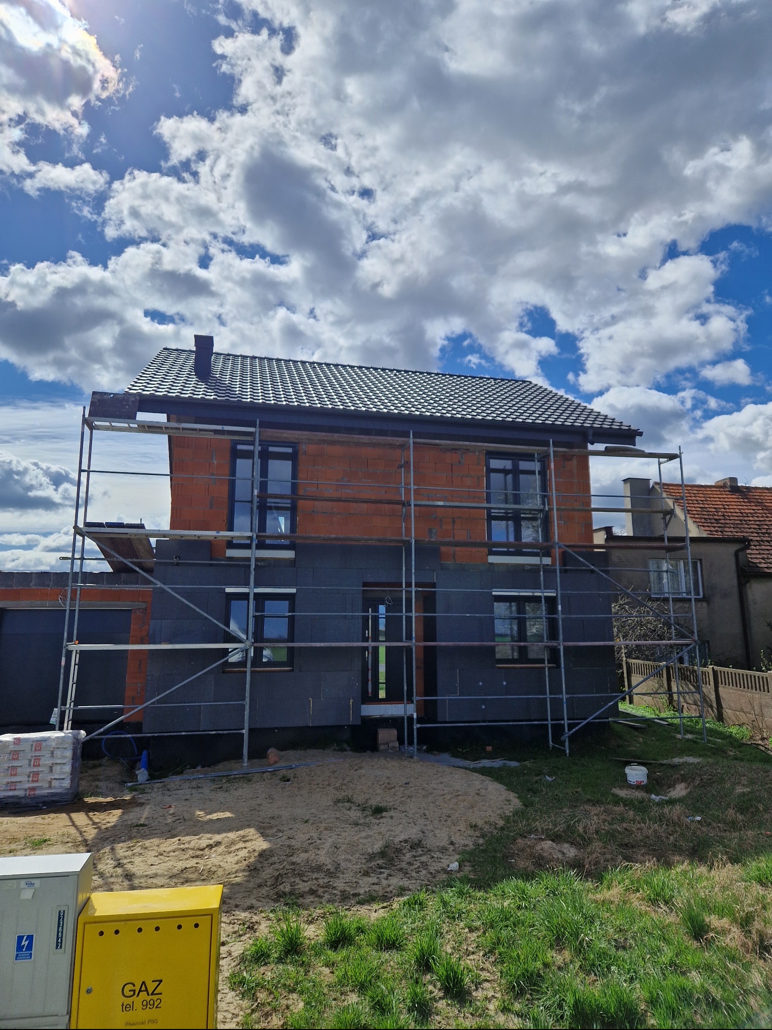 Dom w trakcie budowy, z nowym, ciemnym dachem i rusztowaniem, ocieplany styropianem, na tle błękitnego nieba z chmurami.