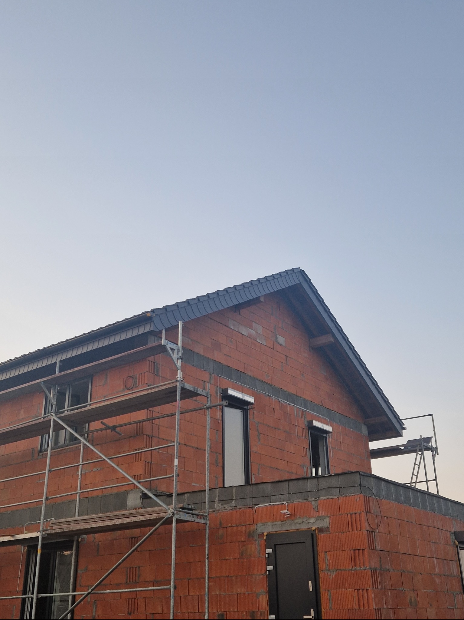 Surowy budynek z czerwonej cegły w trakcie budowy, widoczne rusztowania i fragment dachu z ciemną dachówką, okna częściowo zamontowane, niebo w tle.