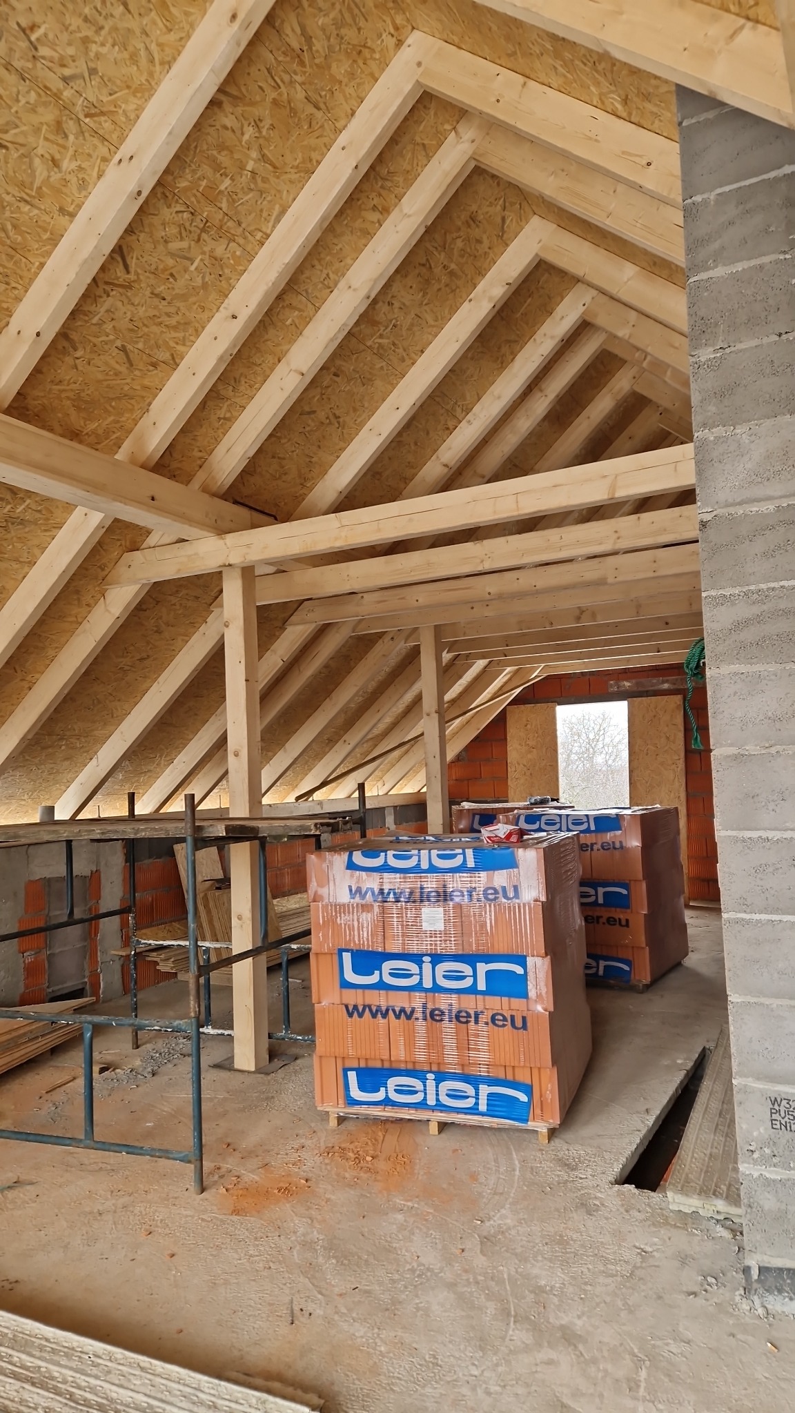 Konstrukcja drewniana dachu z widocznymi krokwiami i deskowaniem OSB, palety z cegłami Leier na budowie.