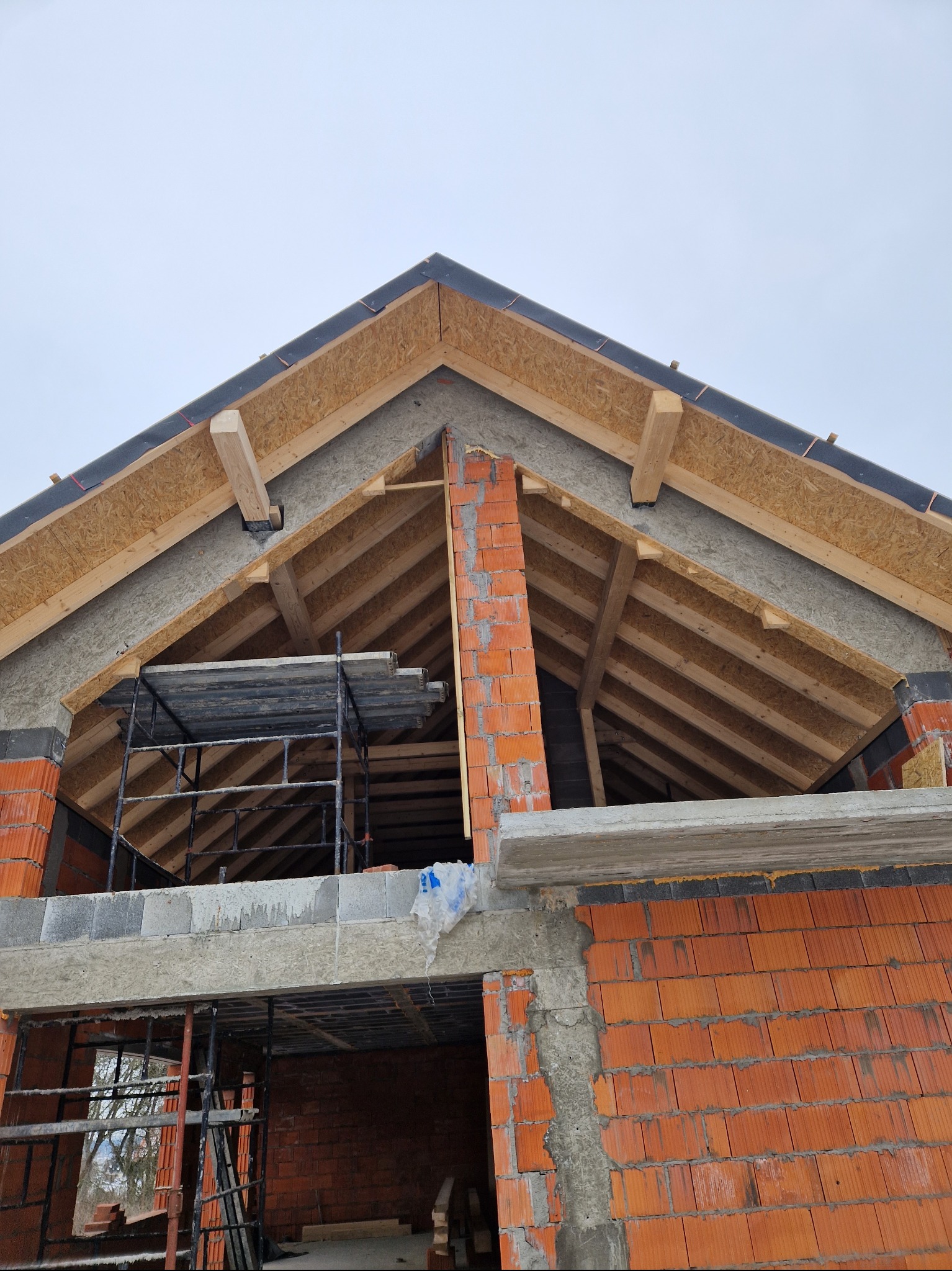 Konstrukcja dachu z widoczną więźbą, deskowaniem, izolacją i kominem z cegły, w trakcie budowy domu z czerwonej cegły, z rusztowaniami w tle.