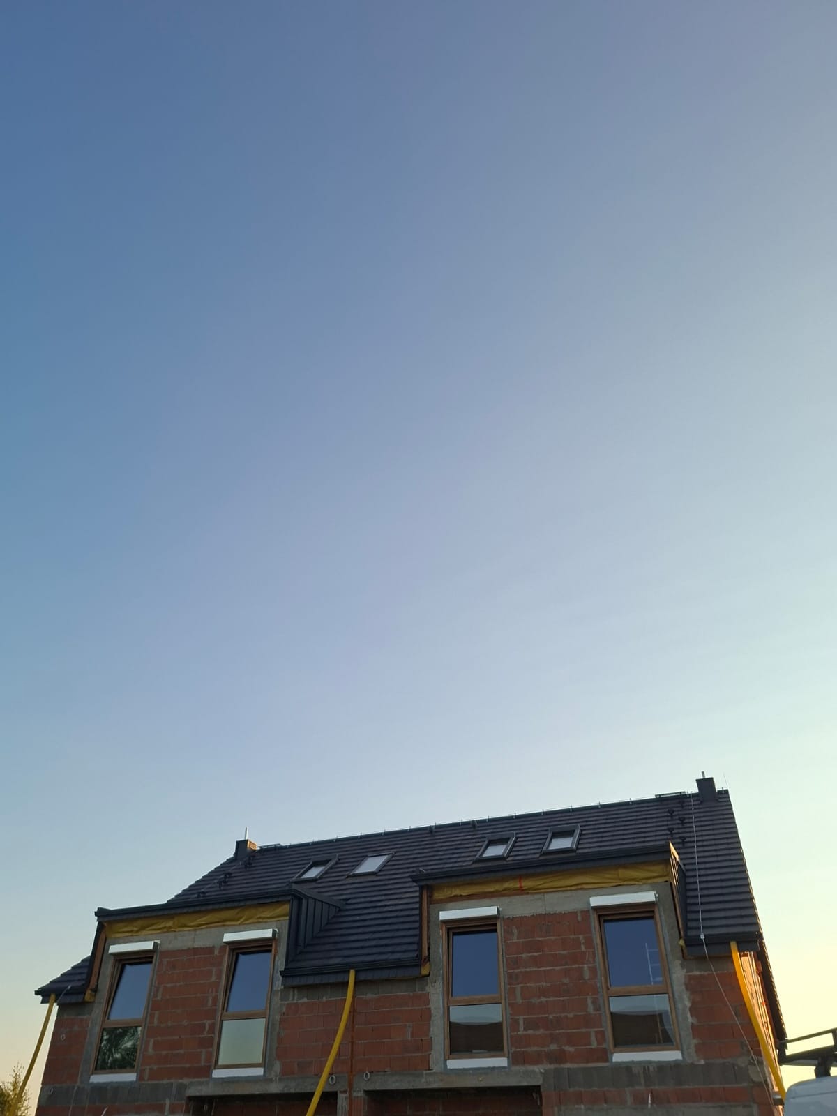 Częściowo wykończony dom z ciemnym dachem i oknami, widoczne pustaki. Żółta folia zabezpieczająca okna. Ujęcie z dołu na tle błękitnego nieba.