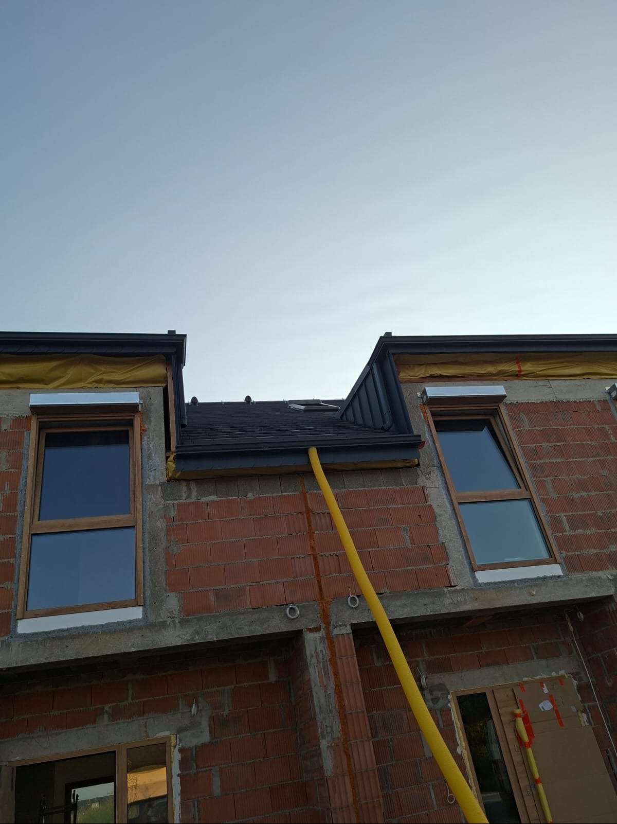 Montaż dachu z blachy na nowym budynku z cegły, widoczne okna dachowe i żółty przewód wentylacyjny. Dach w kolorze grafitowym, wykończenia blacharskie.