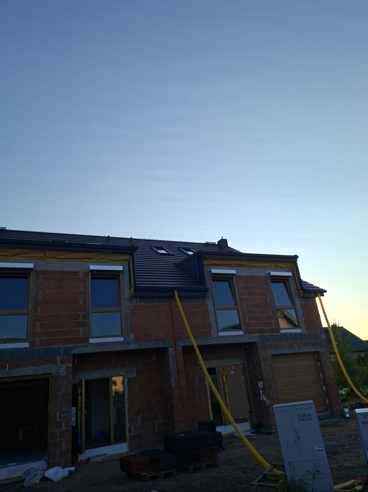 Surowy stan domu w zabudowie szeregowej z oknami i fragmentem dachu. Żółte rury przy elewacji. Widok na niebieskie niebo.