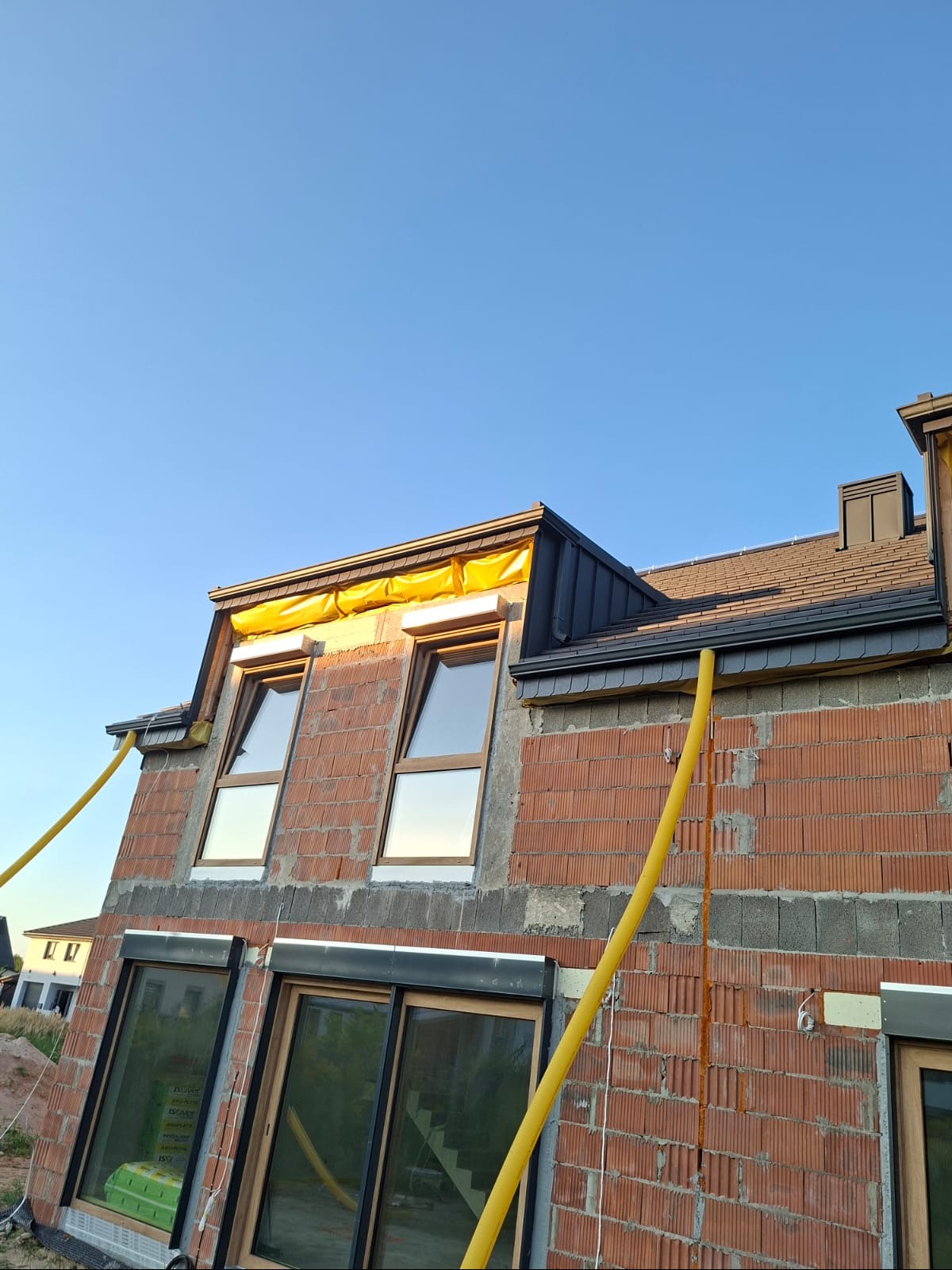 Dom w trakcie budowy: ceglana ściana z oknami, fragment dachu z czarną blachą, żółte rury wentylacyjne. Widok na błękitne niebo.