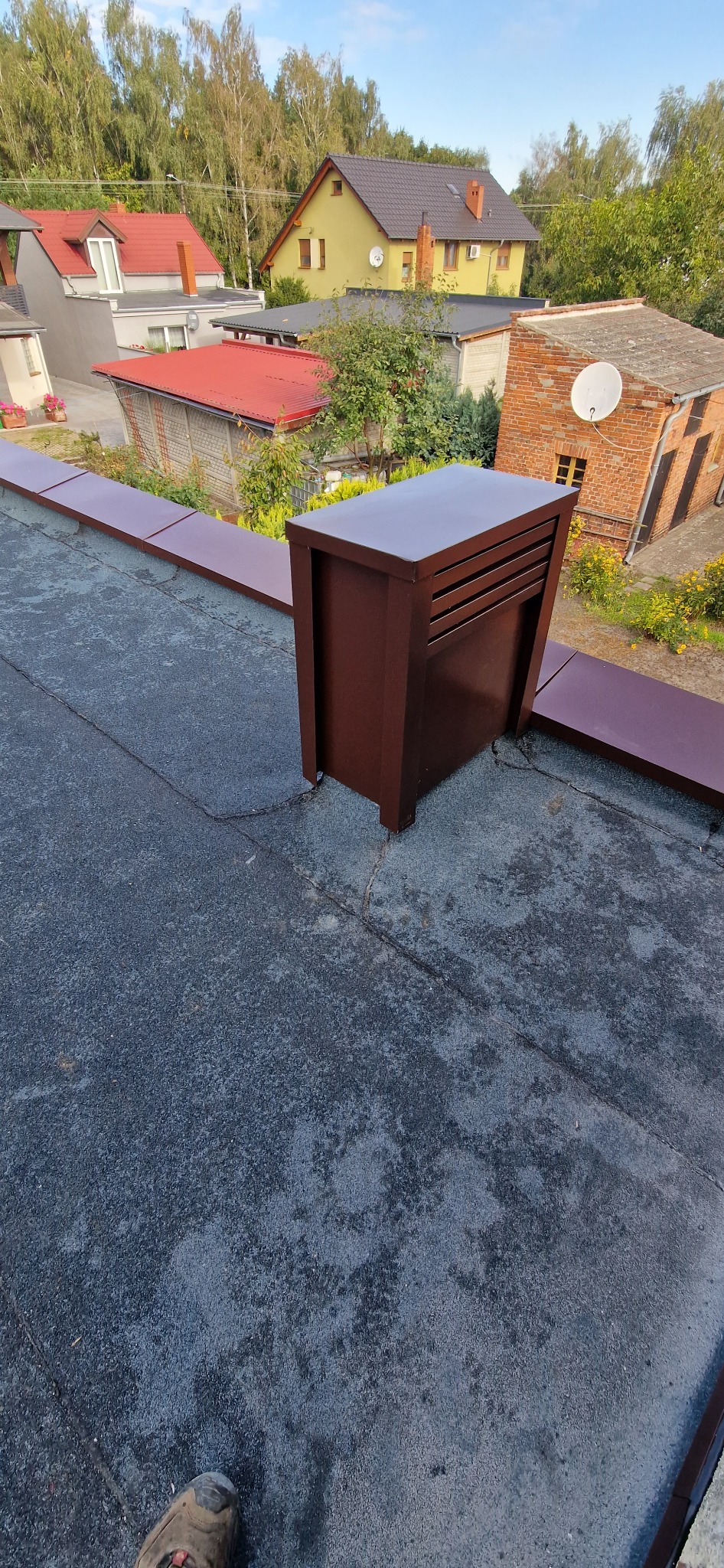 Szczegółowe ujęcie brązowej obróbki blacharskiej komina na dachu pokrytym papą w Krobii, z widocznymi budynkami w tle i fragmentem buta w rogu kadru.