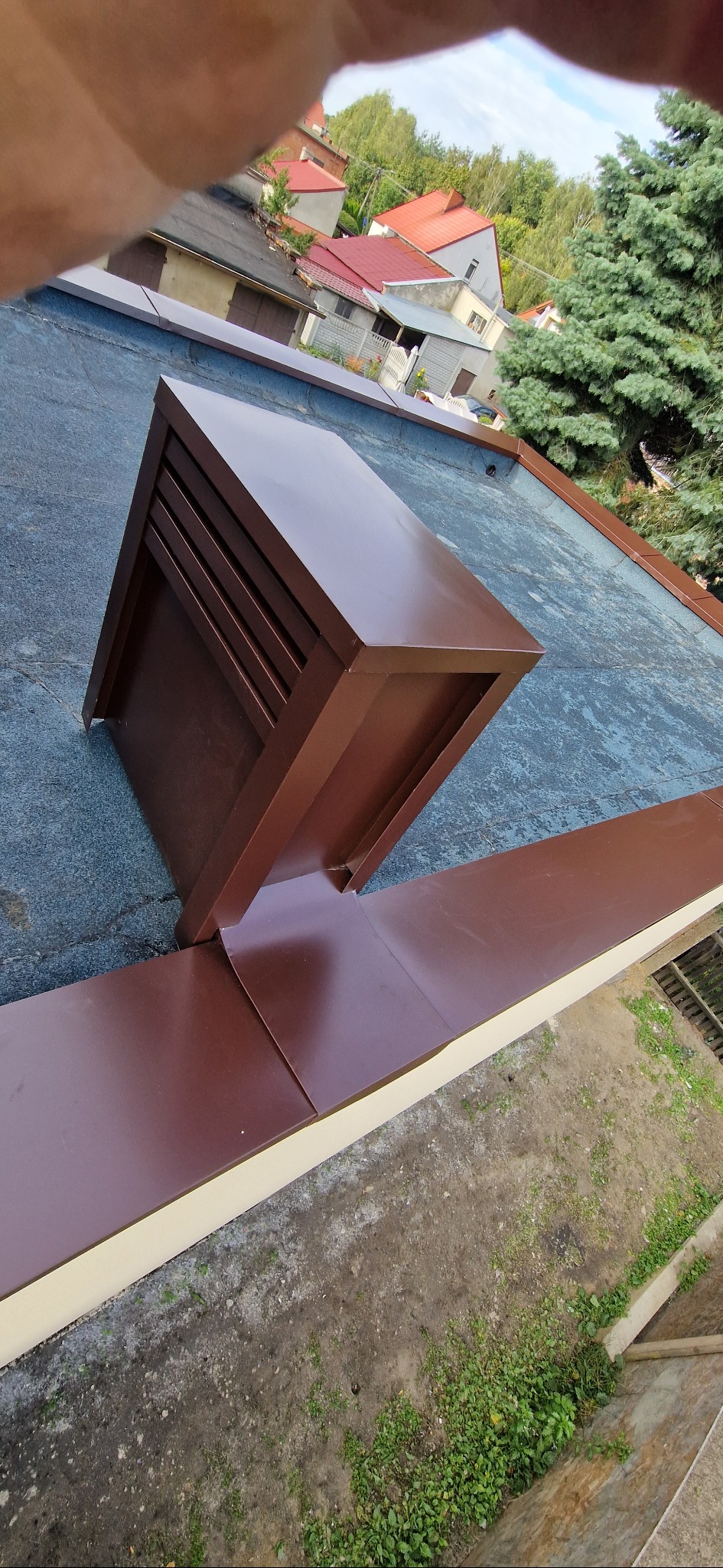 Brązowy kominek wentylacyjny na dachu pokrytym papą, z widocznymi obróbkami blacharskimi i fragmentem zabudowy mieszkalnej w tle.