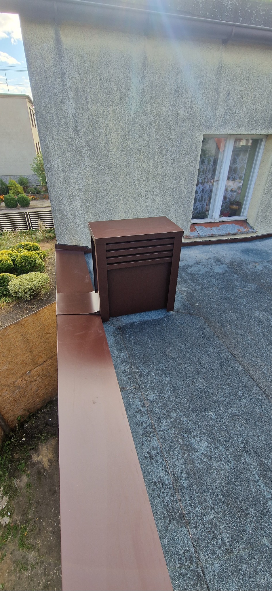 Nowy, brązowy kominek wentylacyjny na dachu z papy, obok obróbka blacharska w tym samym kolorze. W tle budynek z oknem i fragment ogrodu.