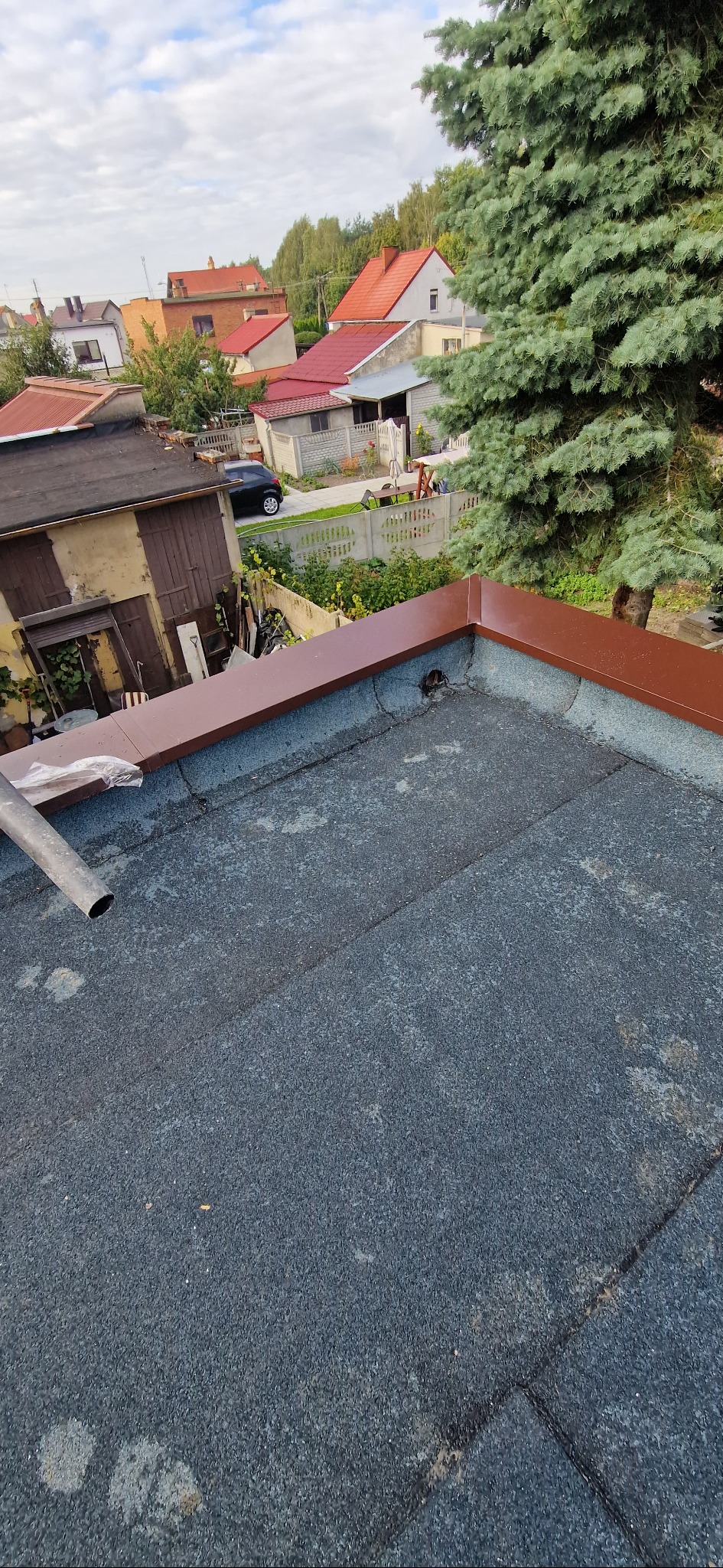 Widok z góry na płaski dach pokryty papą z nowymi, brązowymi obróbkami blacharskimi. W tle zabudowa jednorodzinna i zieleń.