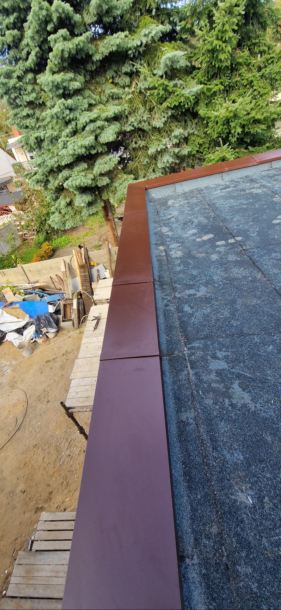 Fragment dachu pokrytego papą z brązowymi obróbkami blacharskimi, widok z góry na tle zieleni drzew. Prace dekarskie w toku, widoczne rusztowania.