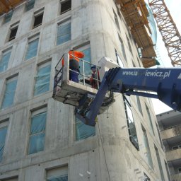 Matpol Budownictwo Sp.zo.o. - Dwóch robotników w pomarańczowej odzieży roboczej na podnośniku koszowym na tle surowego budynku z betonu, w tle widoczny żuraw budowlany.